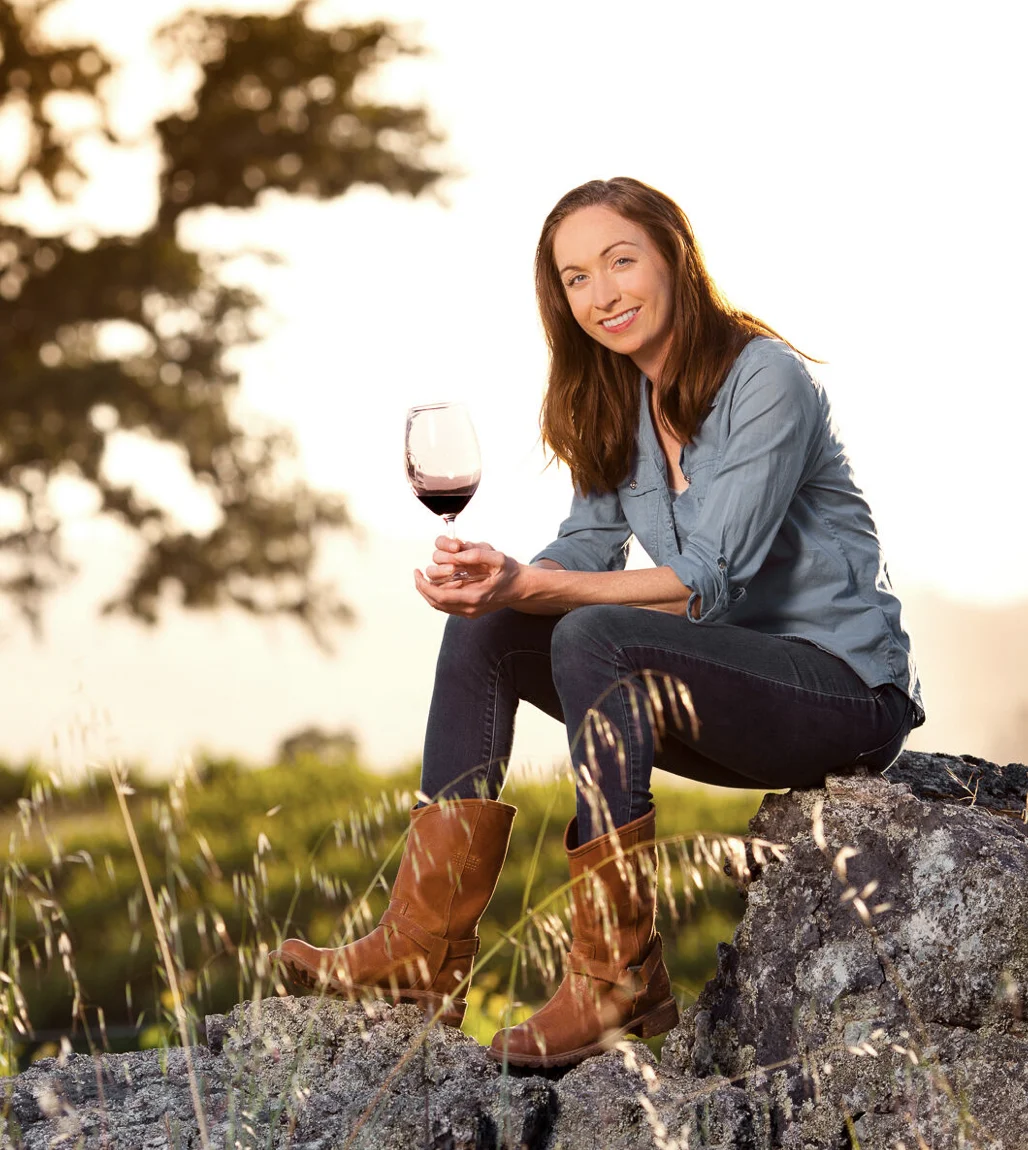 Winemaker 26, a young woman in blue jeans and a blue button up shirt with brown boots sitting on a rock, holding a glass of wine.
