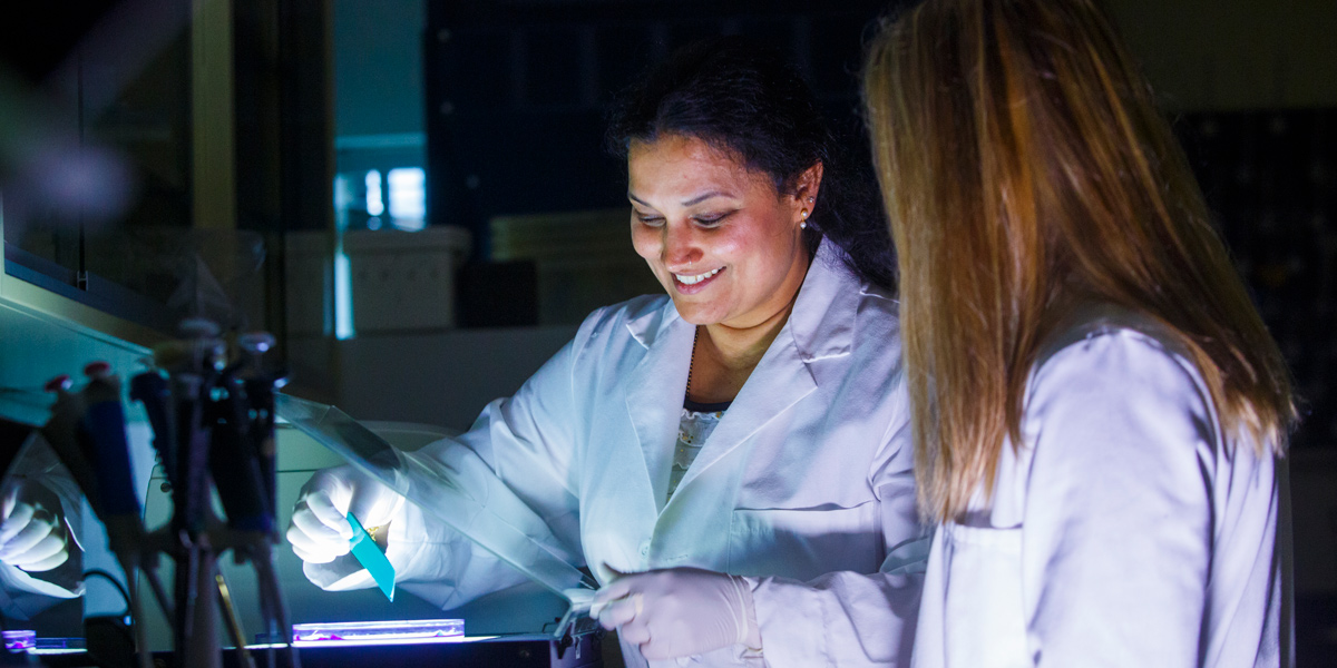 Kucknoor smiling in a lab while showing a female student a specimen underneath a containment shield. Both professor and student are wearing a white lab coat. Ashwini is wearing white gloves.