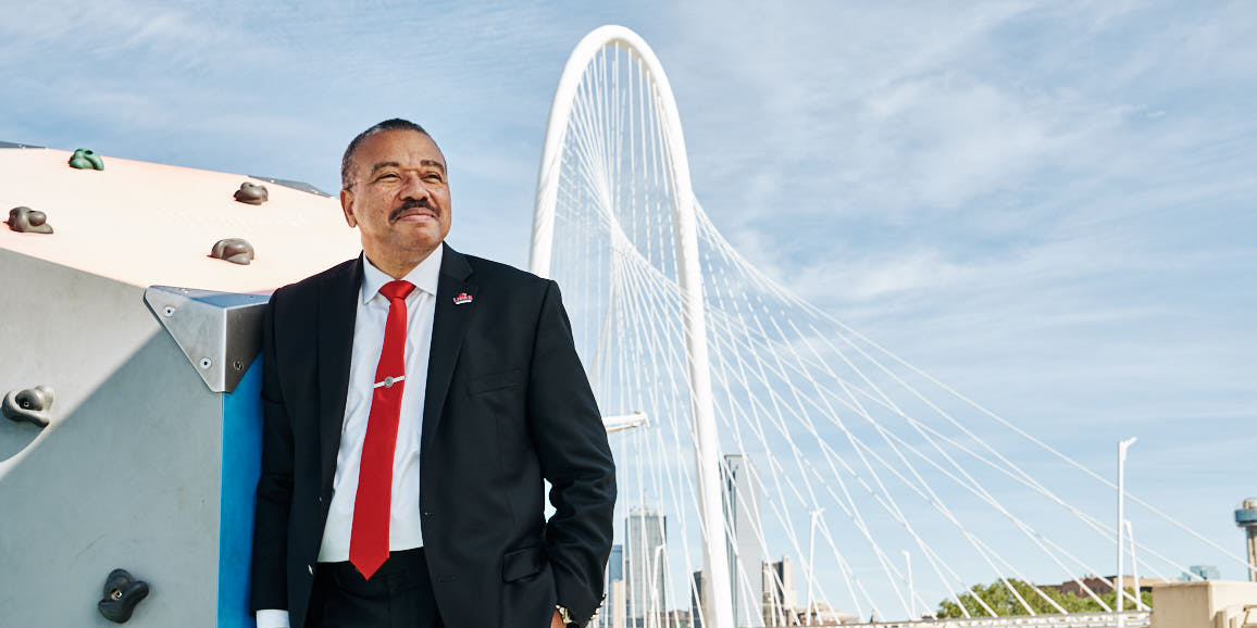 Hyattye Simmons standing in a black suit, hand in pocket. He is wearing a red tie and Lamar pin. Behind him is a large white bridge.