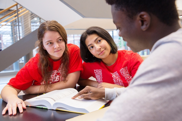 Three students sitting at a table, one student points in a book