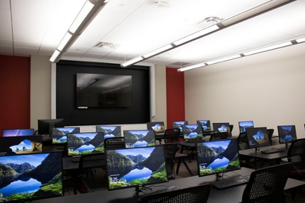 Small room containing several black tables with three computer monitors on each one, white walls and ceiling with a large screen tv, and whiteboards at the back of the room.