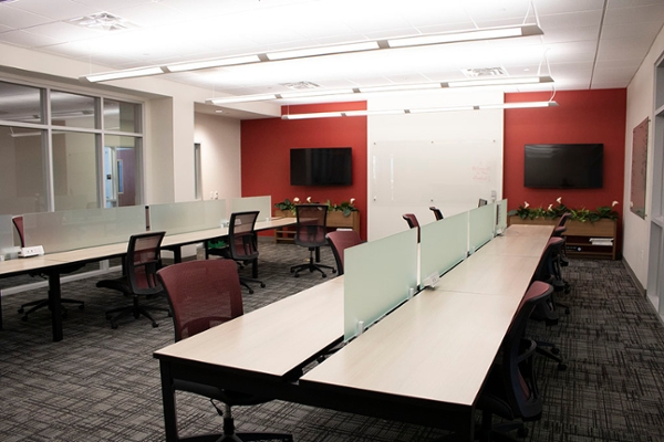 Medium sized room with two rows of wooden tables with central dividers and red chairs. There are two televisions on the back with a whiteboard in the center of the wall, and windows along the side of the room viewing the CICE hallway.