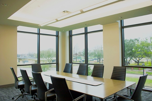 Small, bright room with 10 black office chairs surrounding a large gray table with large paned windows framed by white walls.