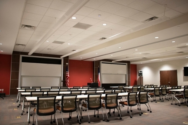 Large room with several rows of white tables and black chairs facing two projector screens on a red wall with a podium in the center.