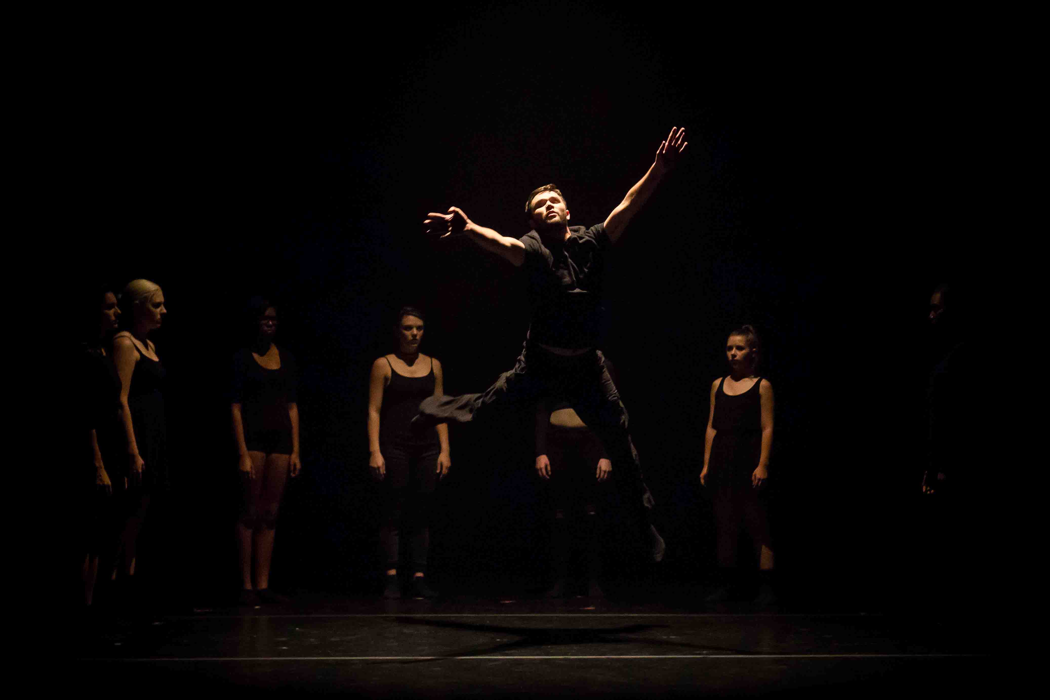 A dancer leaps into the air on a dark stage, surrounded by six standing dancers.