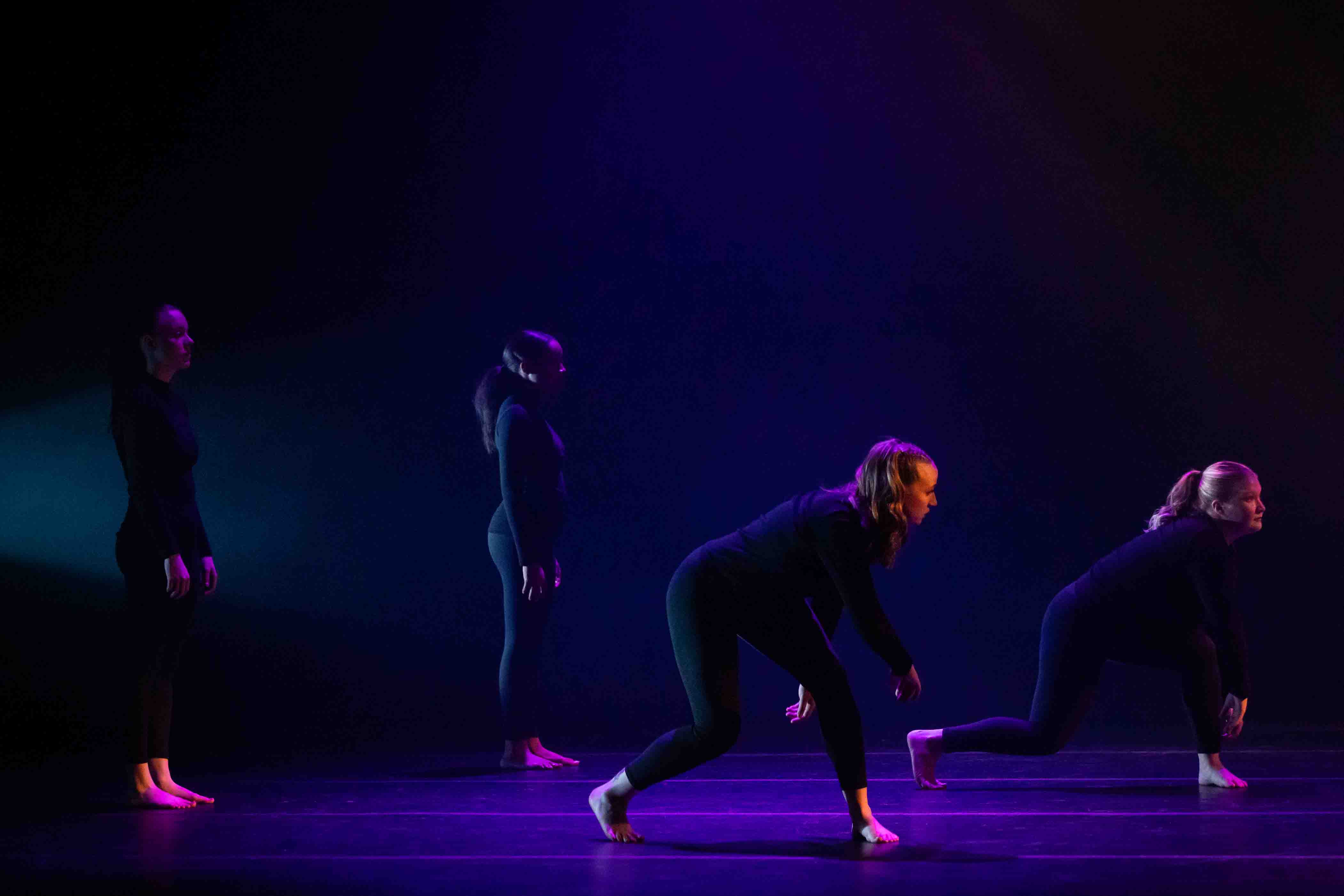 Four dancers in black attire perform on a dimly lit stage with blue and purple lighting.