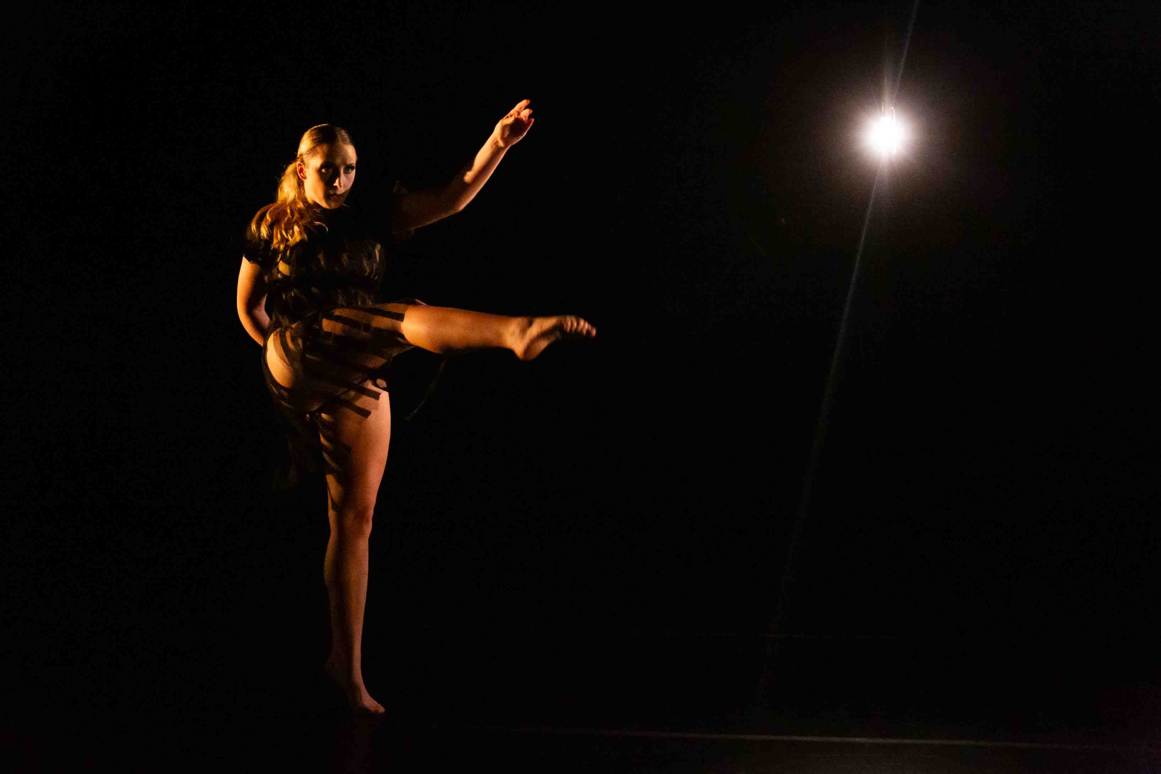 A dancer performs on a dark stage with a spotlight in the background, one leg raised high and arms extended.