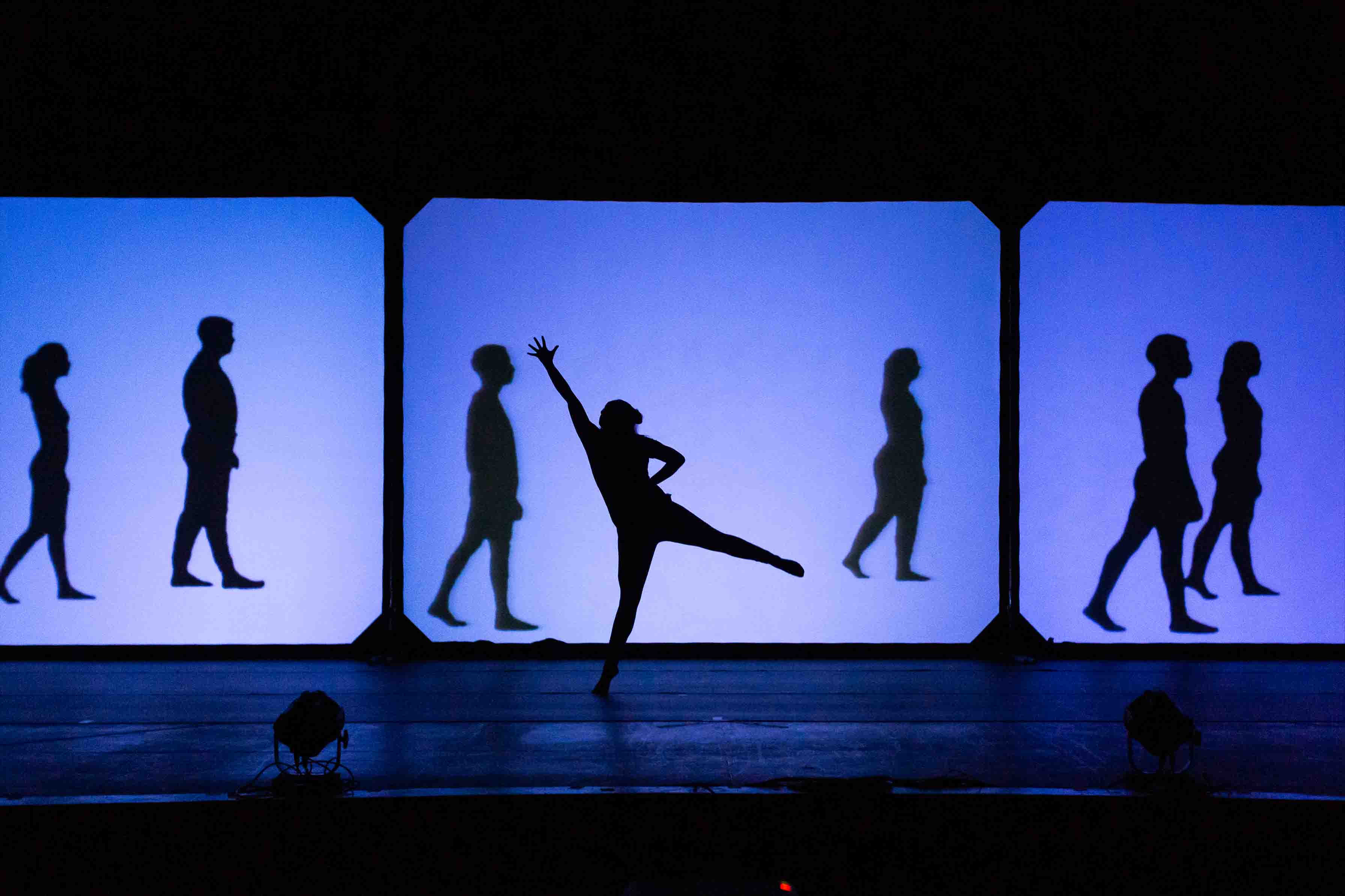 Silhouettes of people walking and a dancer posing on a stage with blue-lit screens.