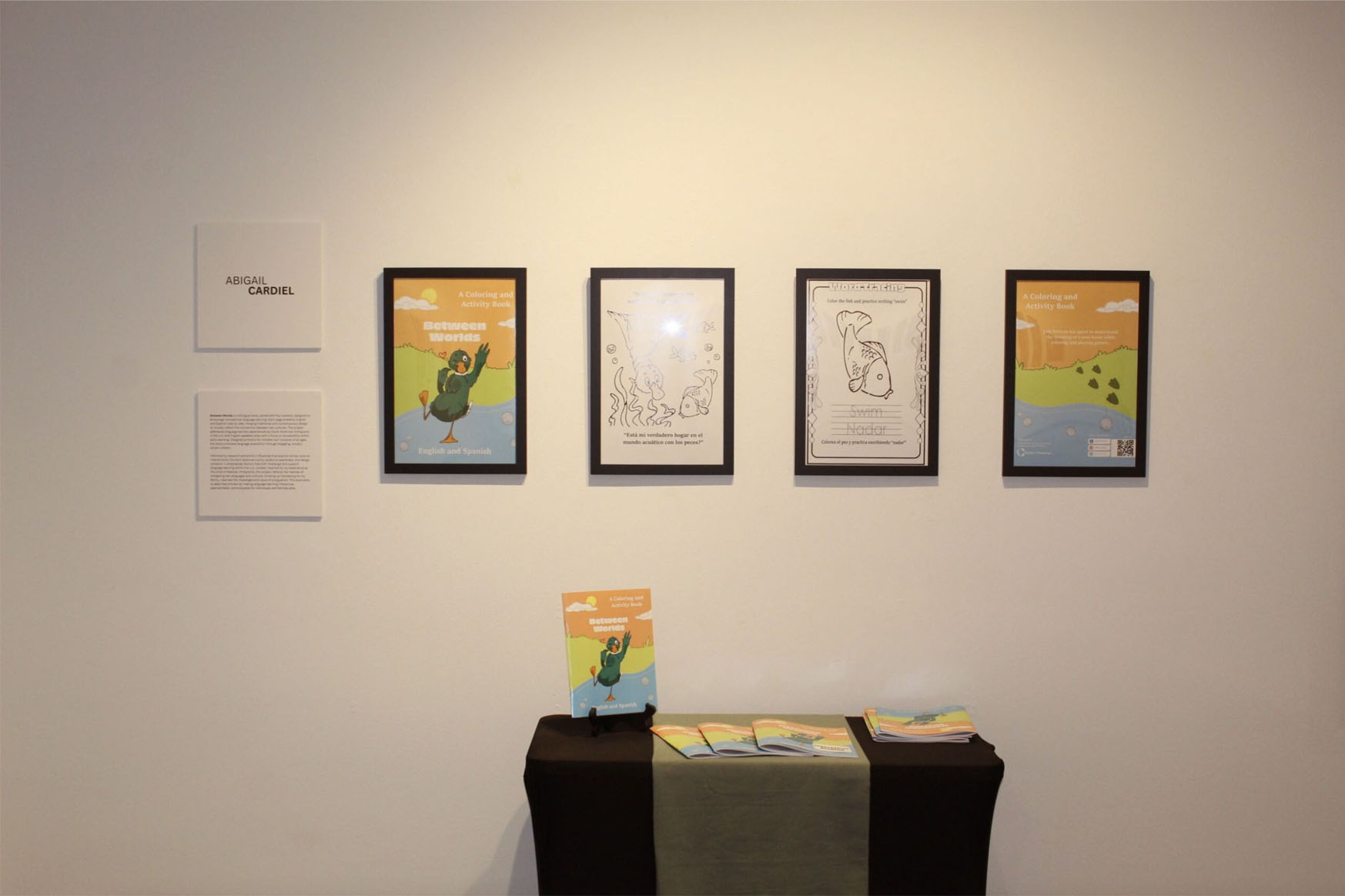 Art display featuring framed artwork and books by Abigail Cardiel, including "Between Worlds" covers and coloring pages.
