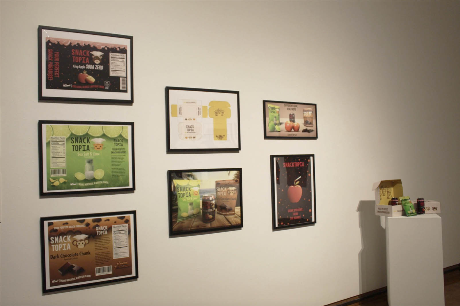 A gallery wall displaying framed product packaging designs for Snack Topia and a pedestal with snack items.