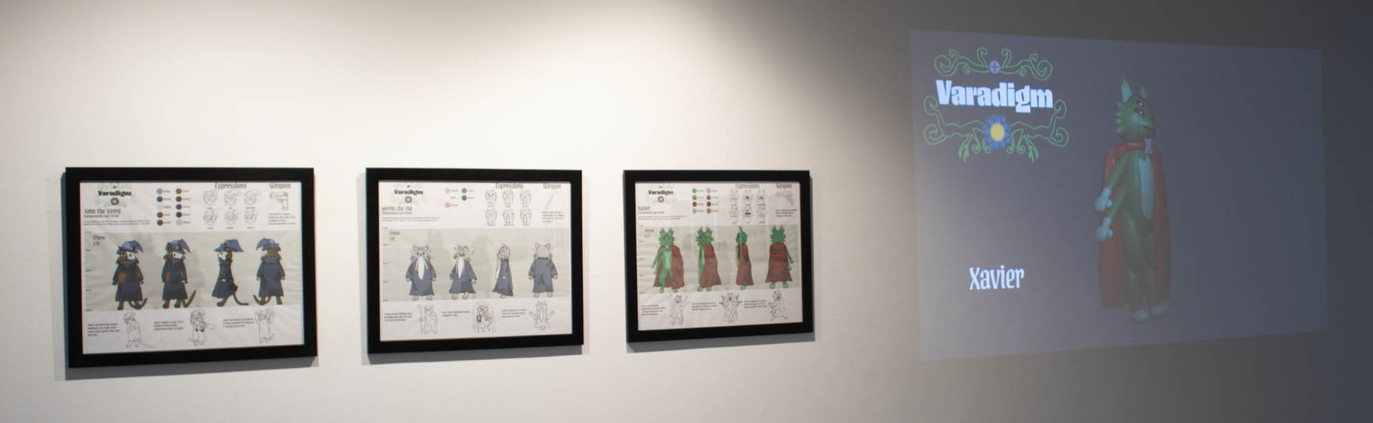 Three framed character design artworks and a projection of a green humanoid creature named Xavier with a red cape, alongside the title "Varadigm."