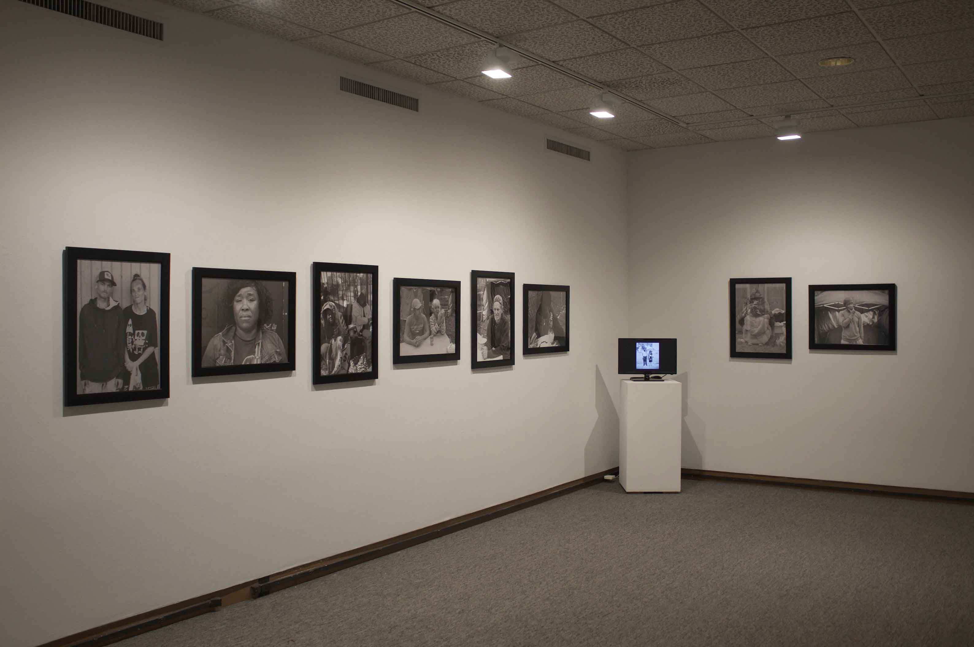An art gallery with black-and-white photographs on white walls, and a small monitor on a pedestal displaying an image.