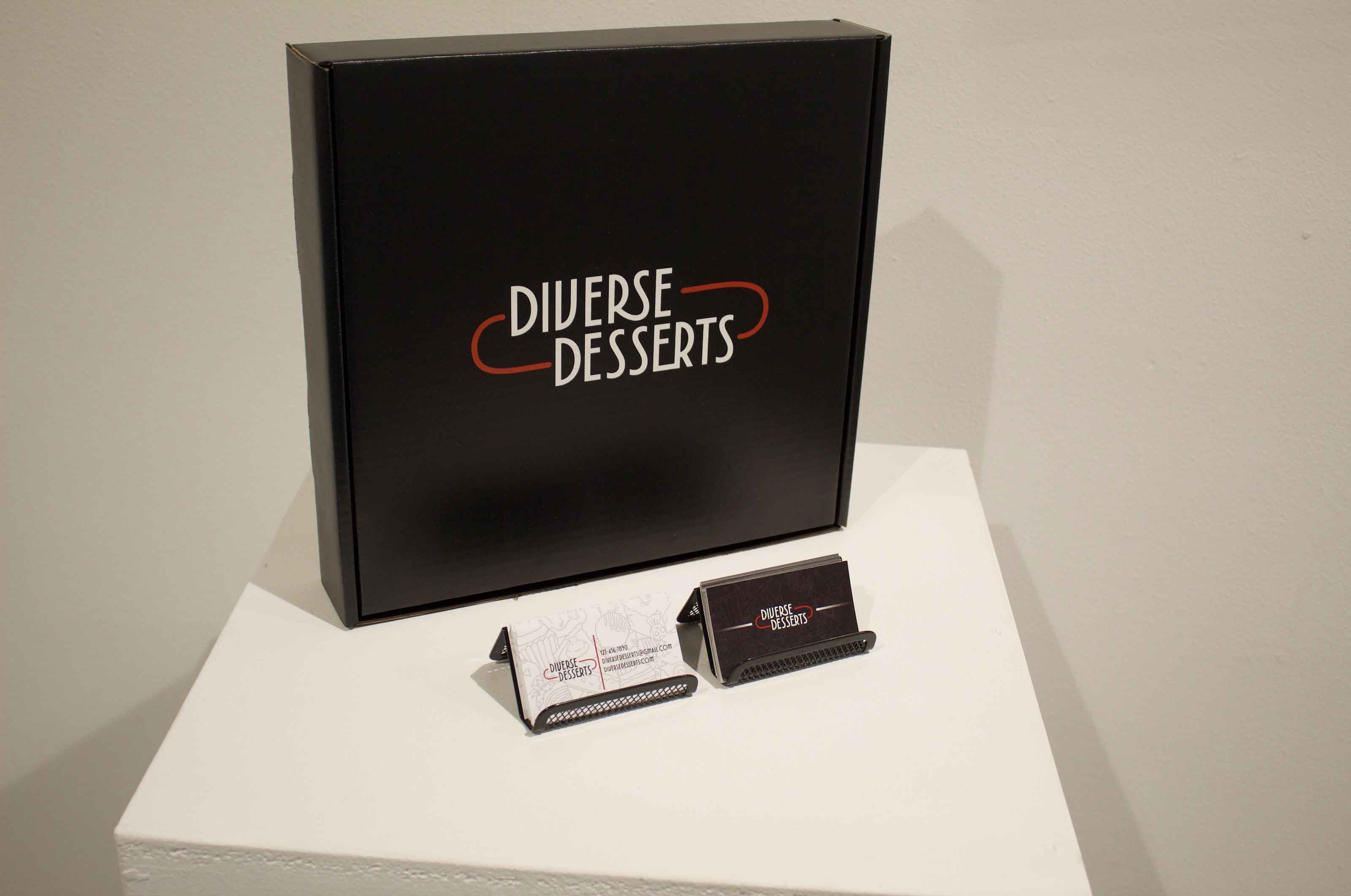 Black box with "DIVERSE DESSERTS" text beside business cards on a white pedestal.