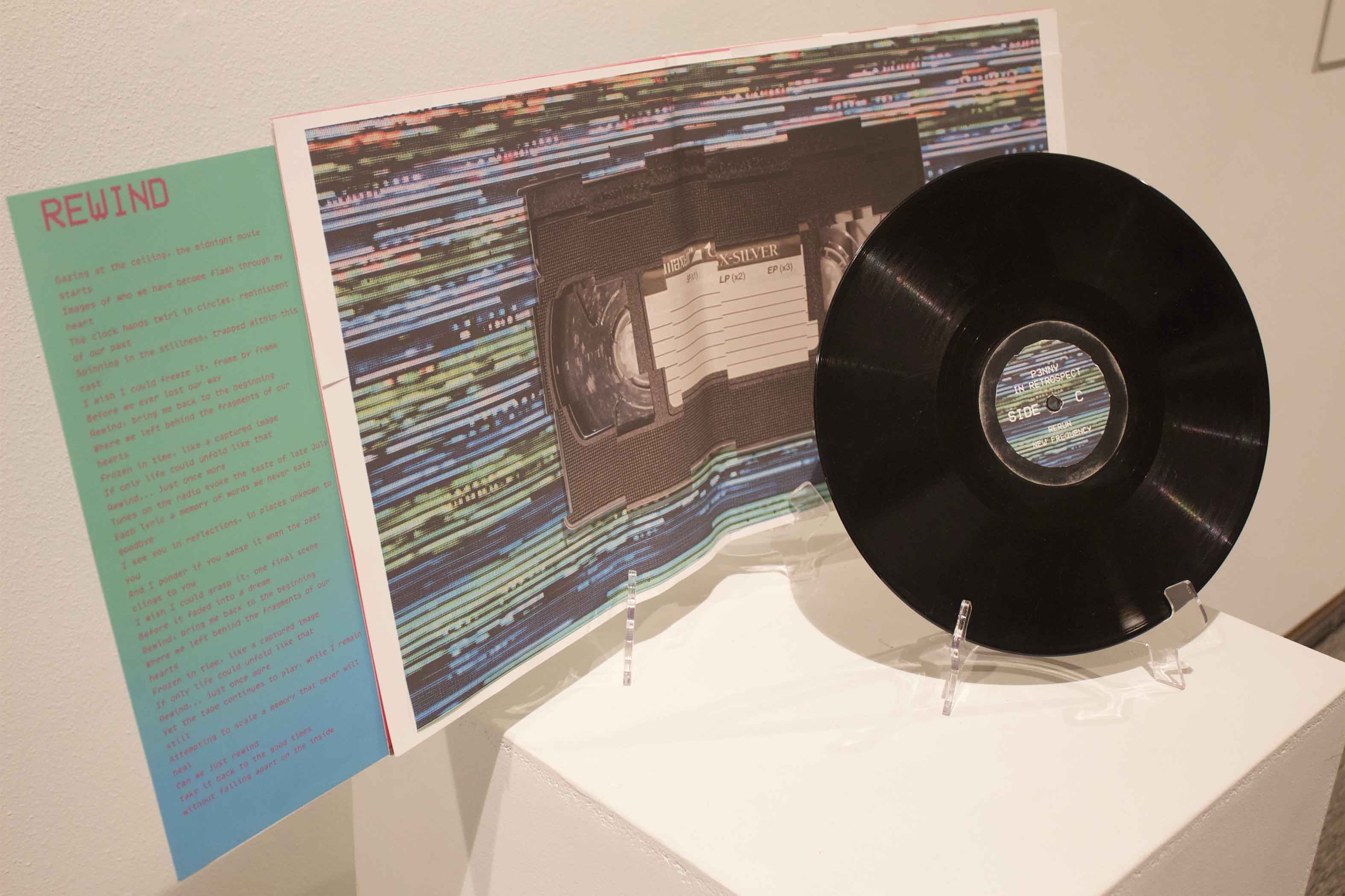 An art installation with a VHS tape poster, a black vinyl record, and teal paper with text titled "REWIND."