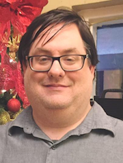 Cris I. Nunn - Taken indoors with partial holiday decorations in the background. Cris, wearing a casual grey colored button down shirt and black framed eyeglasses, is smiling at the camera.