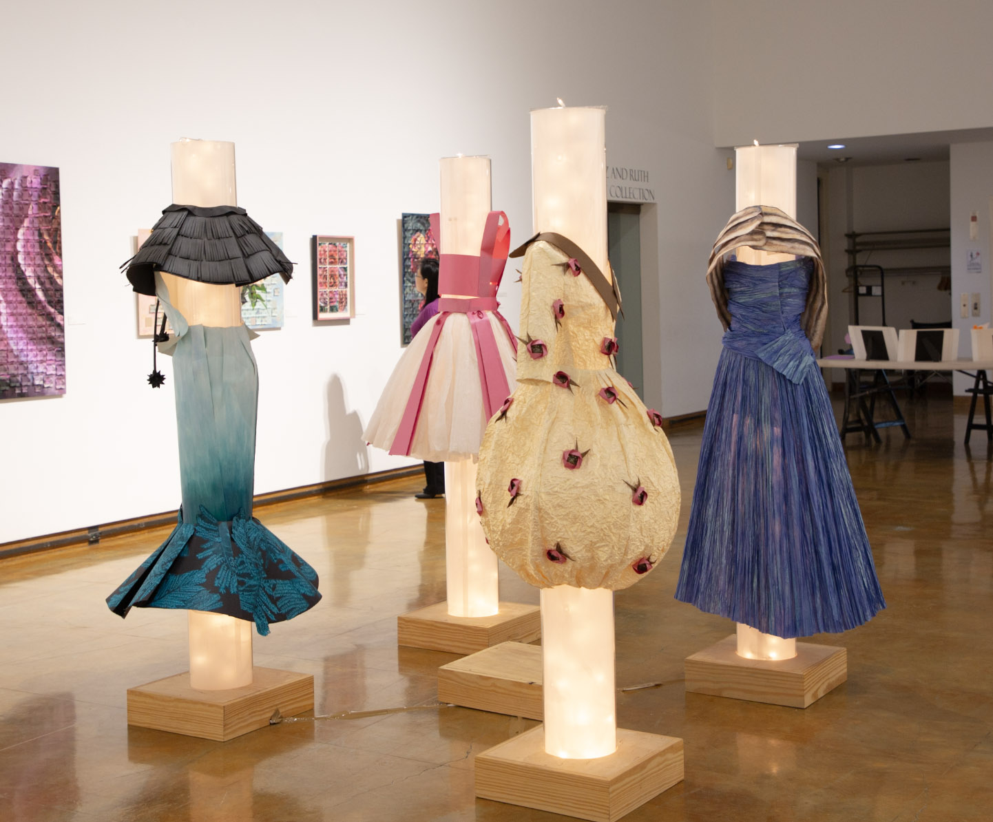 Artistic dress forms displayed on illuminated pedestals in a gallery.