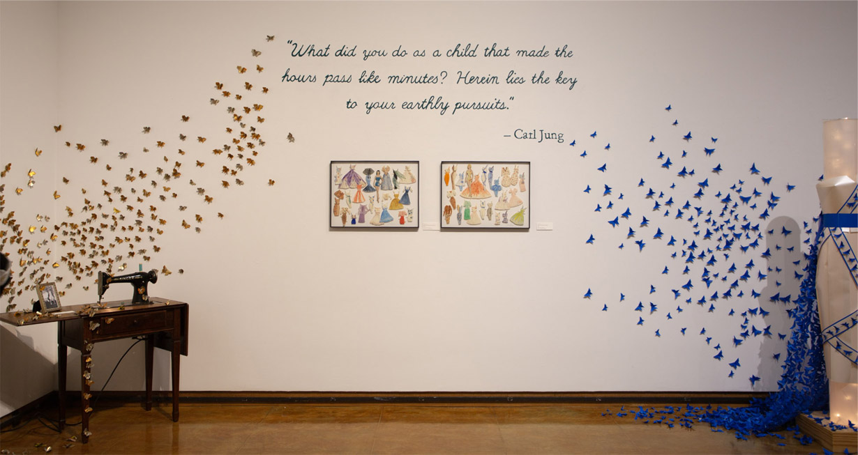 A wall art installation with golden butterflies and blue origami birds, a sewing machine, collages of dress designs, and the Carl Jung quote, What did you do as a child that made the hours pass like minutes? Herein lies the key to your earthly pursuits.