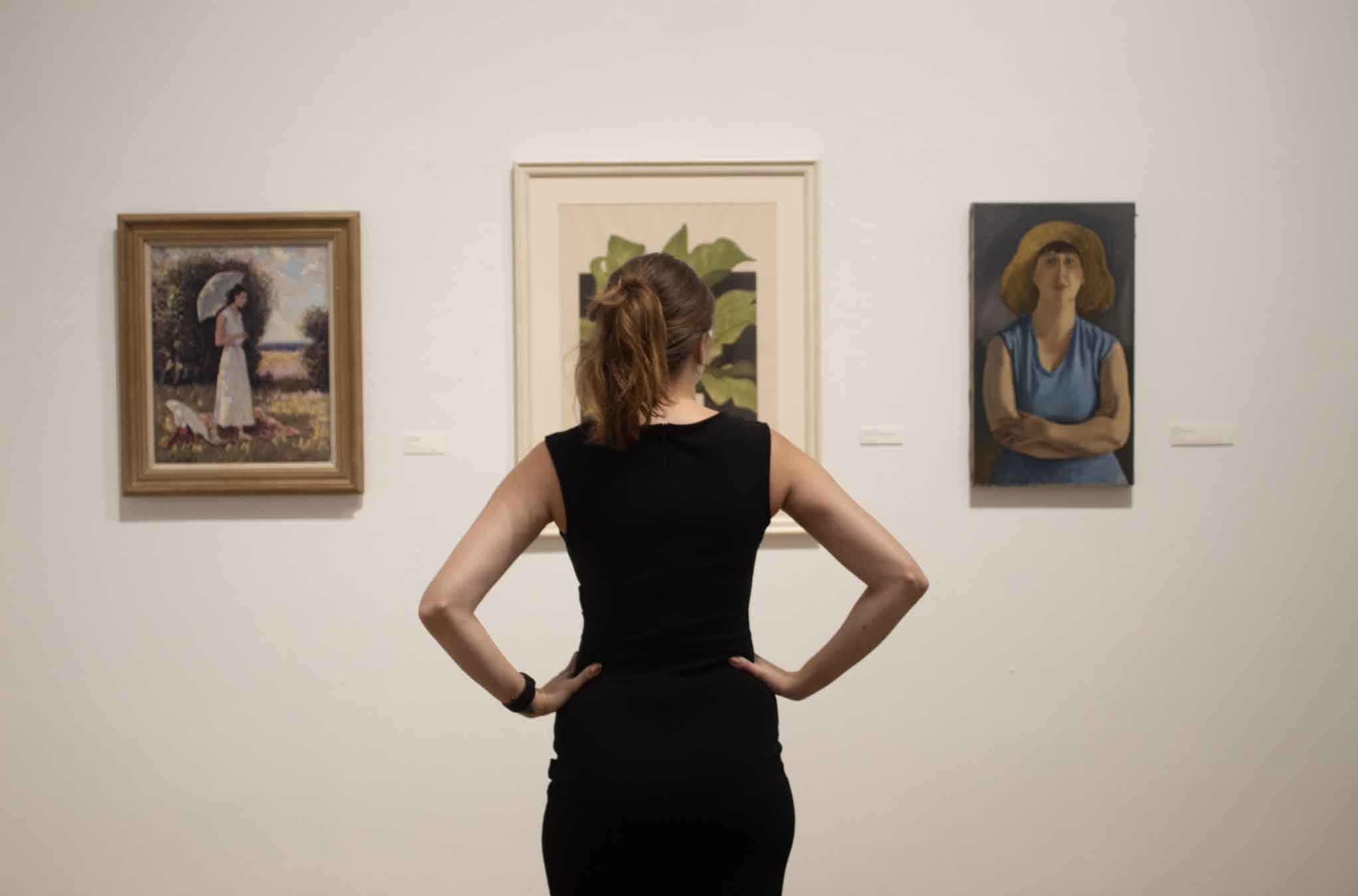 Person in a black dress observing three paintings in an art gallery.