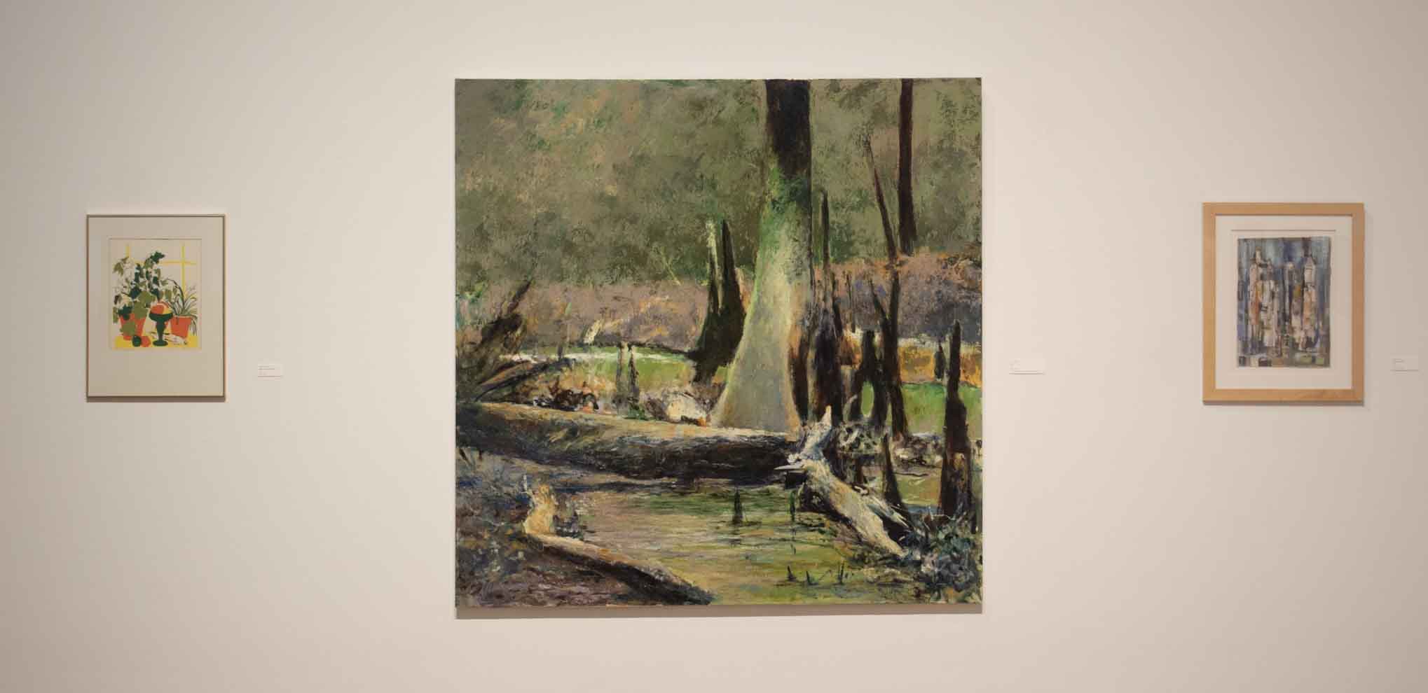 Three framed artworks on a gallery wall: a central large painting of a forest scene flanked by smaller colorful and abstract images.