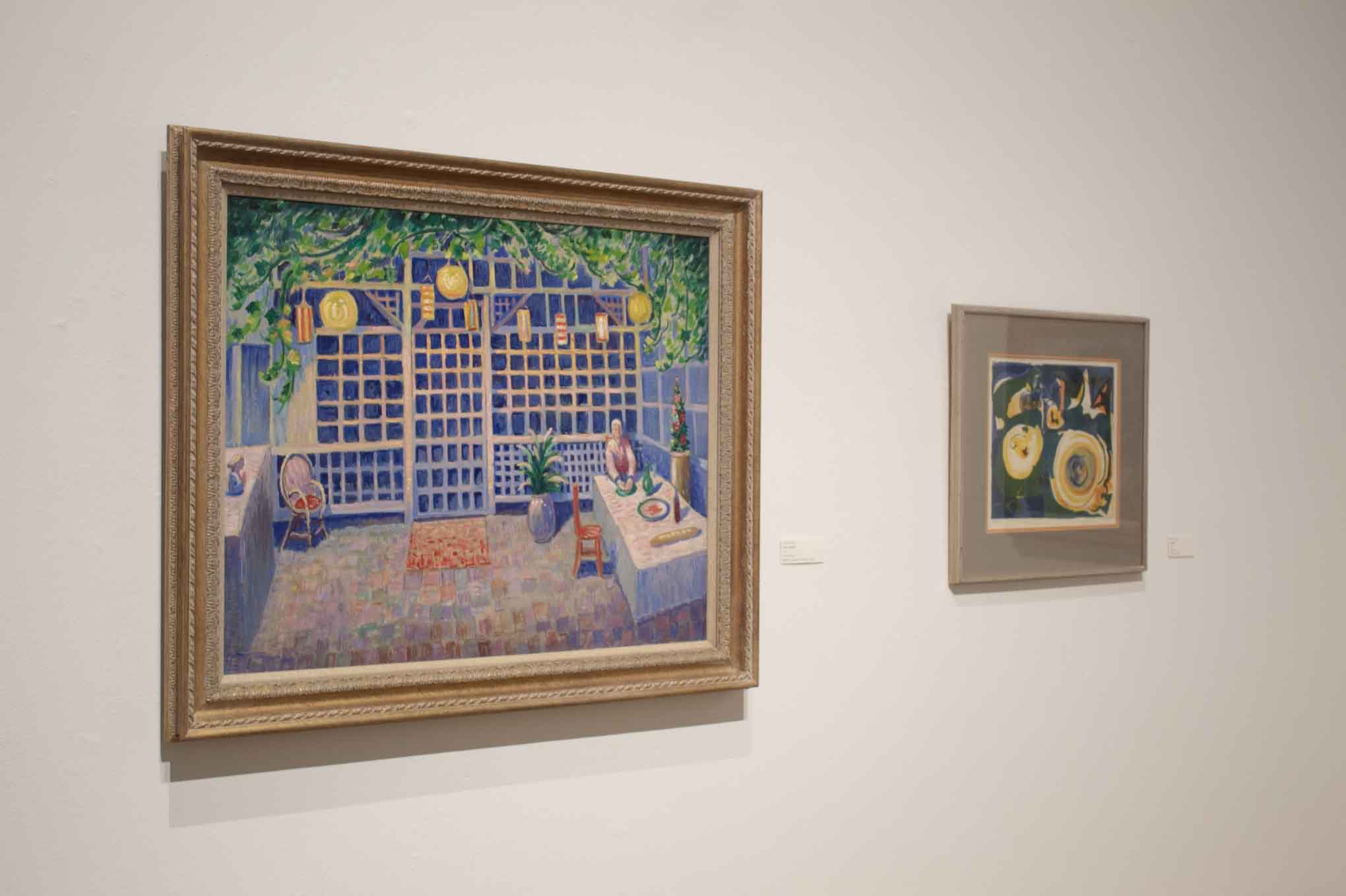 Two framed paintings on a gallery wall: one depicts a vibrant interior scene, the other is abstract with swirling shapes.