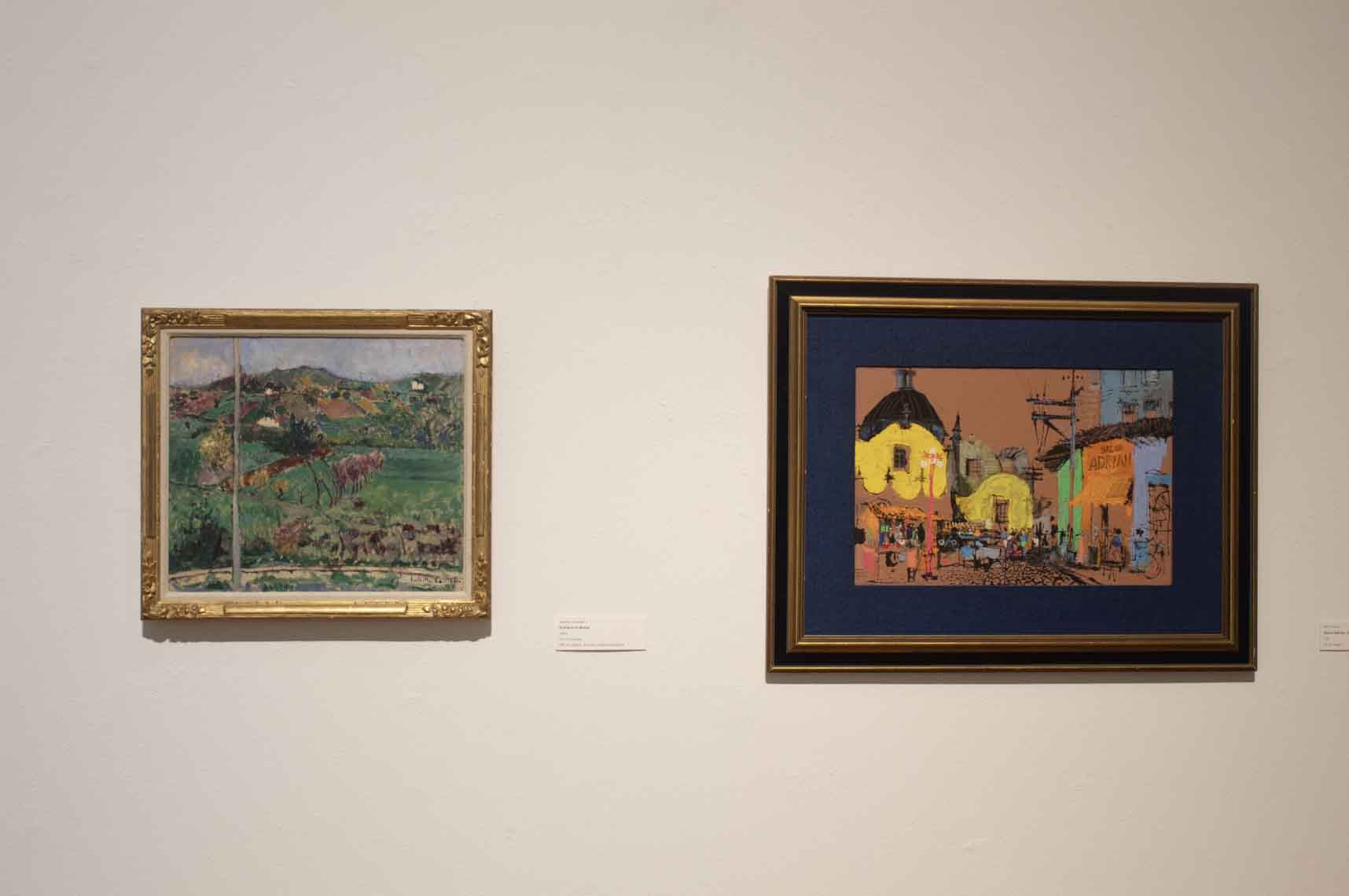 Two framed artworks on a gallery wall, one landscape and one vibrant street scene.