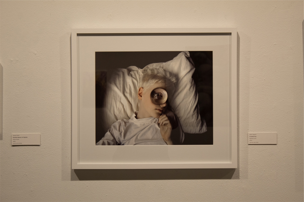 A close-up photograph of a young person lying on a bed, holding a large magnifying glass over one eye, featuring a soft and neutral background.