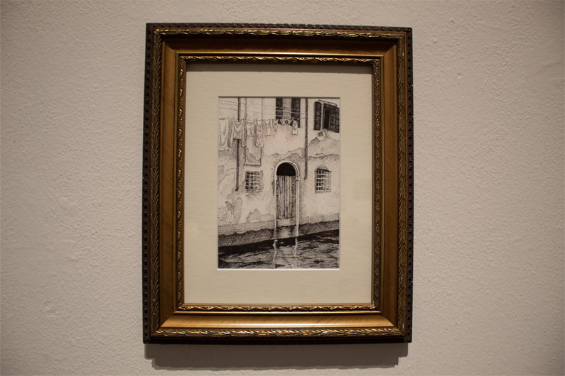 A framed artwork depicting a weathered wooden door opening into a Venetian canal with window openings and hanging laundry.