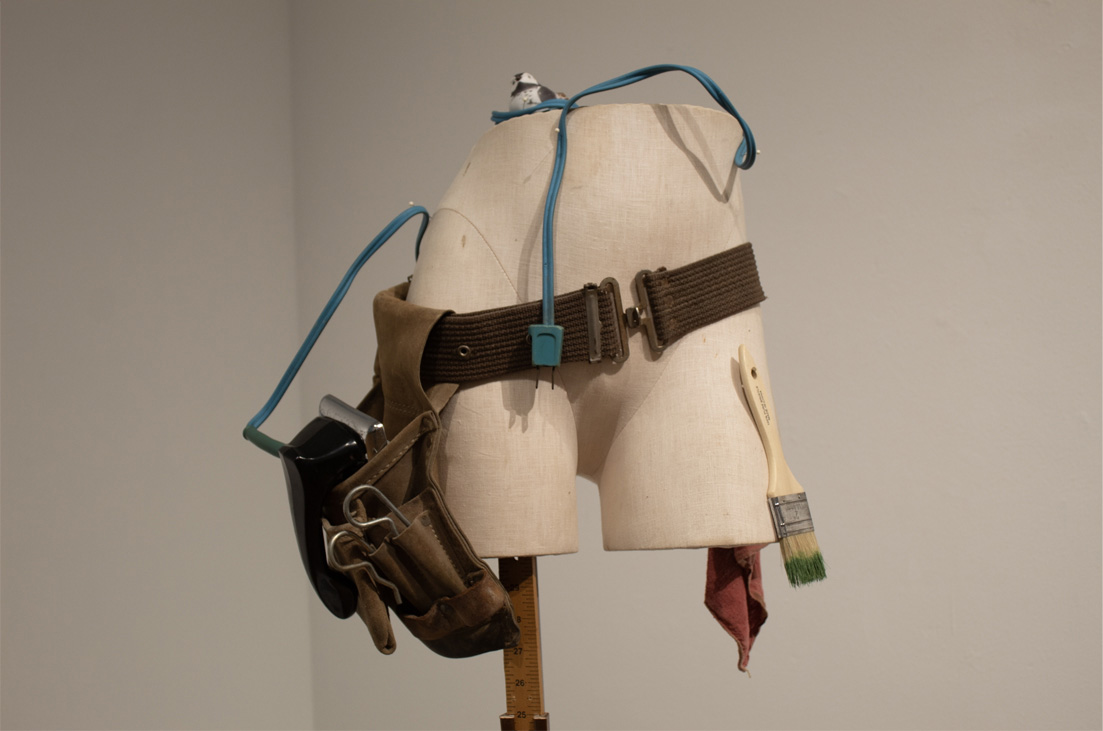 A mannequin torso displays a brown utility belt with a tool pouch, a compact black object, a paintbrush, and a red rag, set against a simple background.