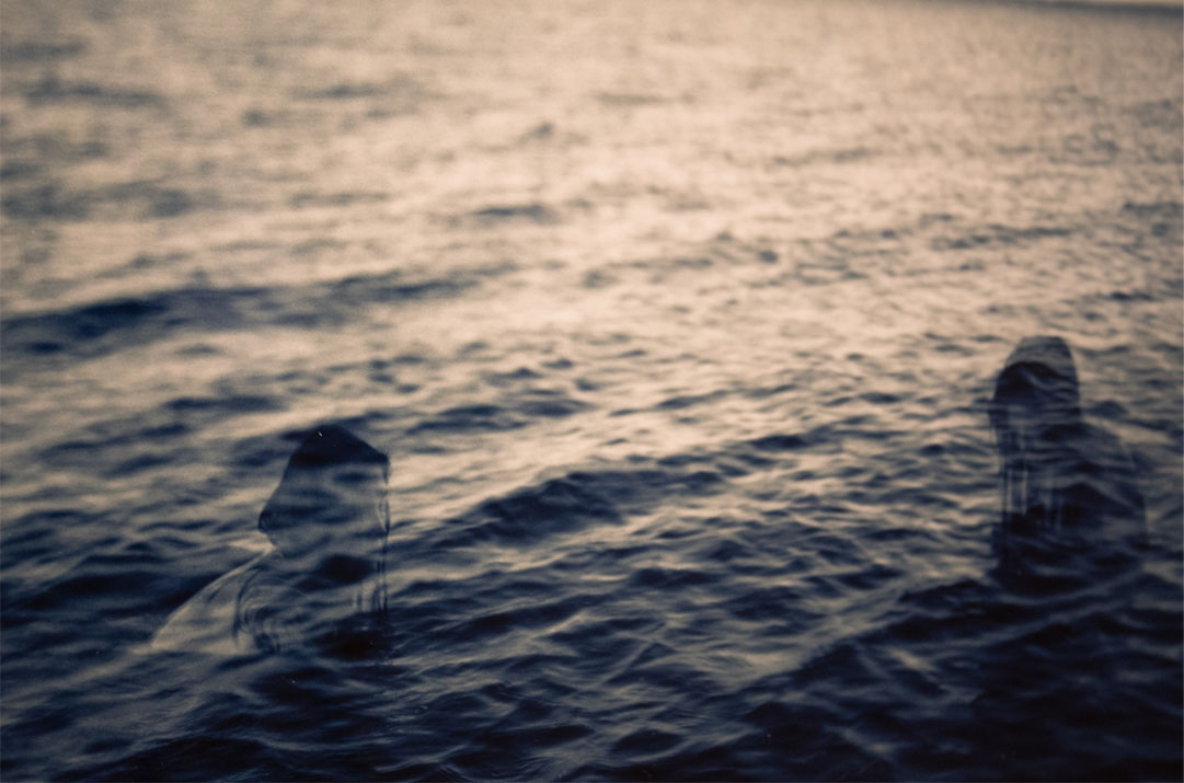 Two indistinct figures partially submerged in dark, textured water, creating a ghostly effect against a blurred background.