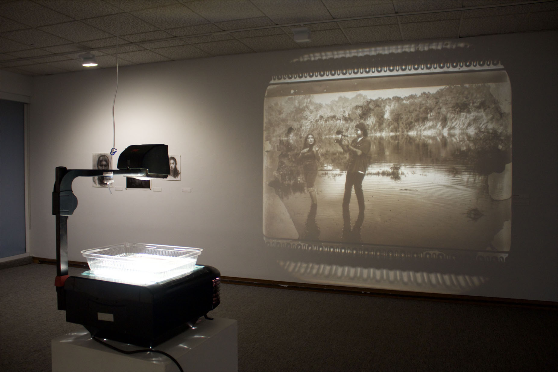 An overhead projector casts a sepia-tone image of two people in a natural setting onto a wall in a dimly lit room.