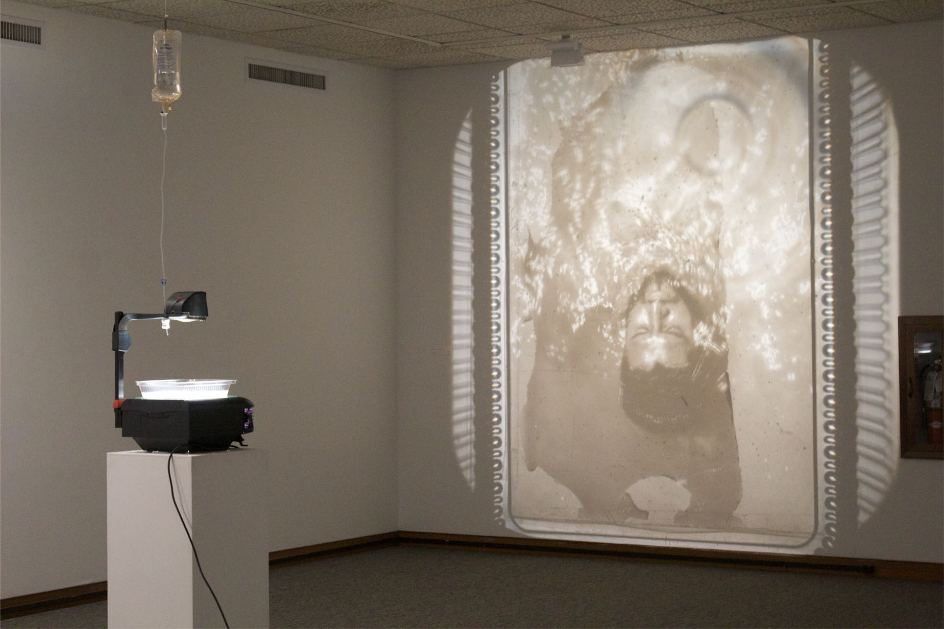 A projector displays a sepia-toned image of an inverted submerged figure onto a wall.