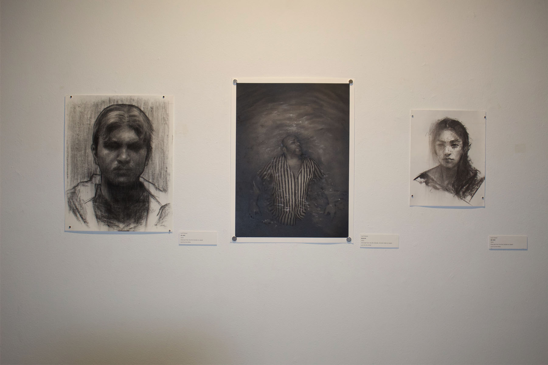 Three monochromatic artworks on a gallery wall; two charcoal portraits and a central photograph of a man in water.