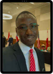Tajudeen Babayeju, smiling man with mustache and glasses wearing dark suit, white shirt, red stripped tie