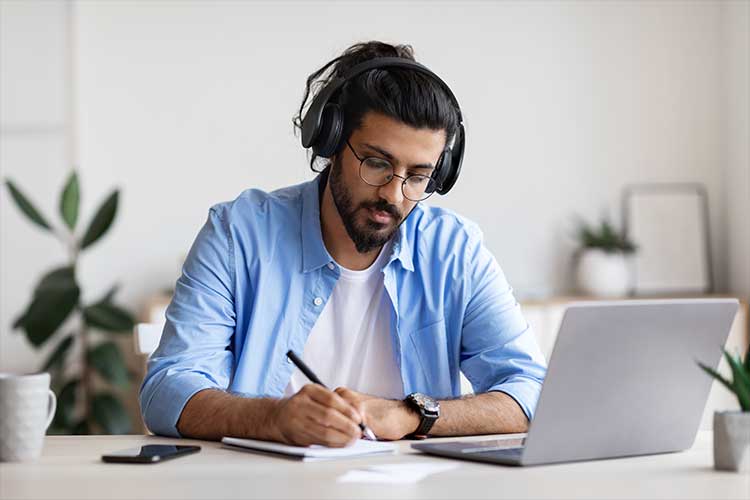 Image shows a young man with dark hair, a beard, and glasses, taking notes in front of an open laptop.